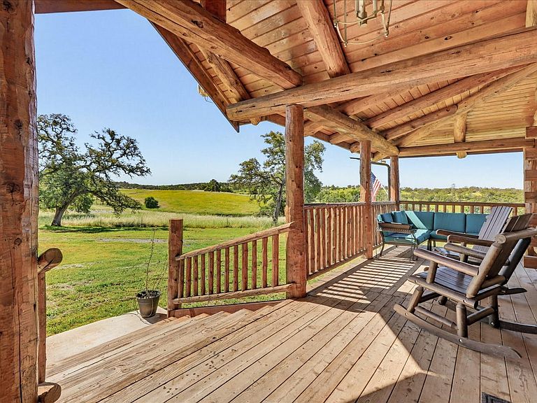Wraparound porch of 20012 White Oak Dr looking out across green rolling pasture toward distant oak-studded ridges, with a turquoise sectional sofa, exposed log beams, and an American flag in afternoon light