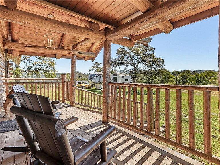 Hand-hewn log porch railings frame a pair of dark wood Adirondack rockers, with a fifth-wheel RV parked on the ranch beyond and oak trees scattered across the pasture
