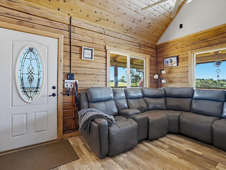Interior of the log cabin great room with a vaulted tongue-and-groove pine ceiling, large picture windows looking onto the pasture, and a leather sectional facing the entry