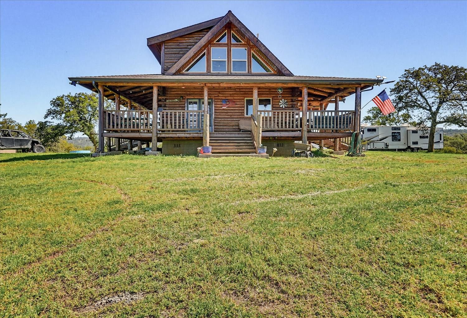 Front elevation of a two-story log cabin at 20012 White Oak Dr, with a wraparound porch, American flag, and oak-studded rolling pasture under a wide blue California sky